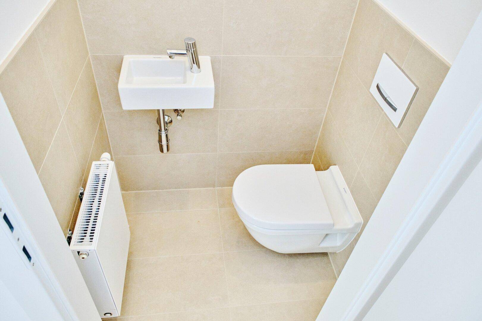 Kompaktes Gäste-WC mit wandhängender Toilette, kleinem Waschbecken und beigefarbenen Wand- und Bodenfliesen.