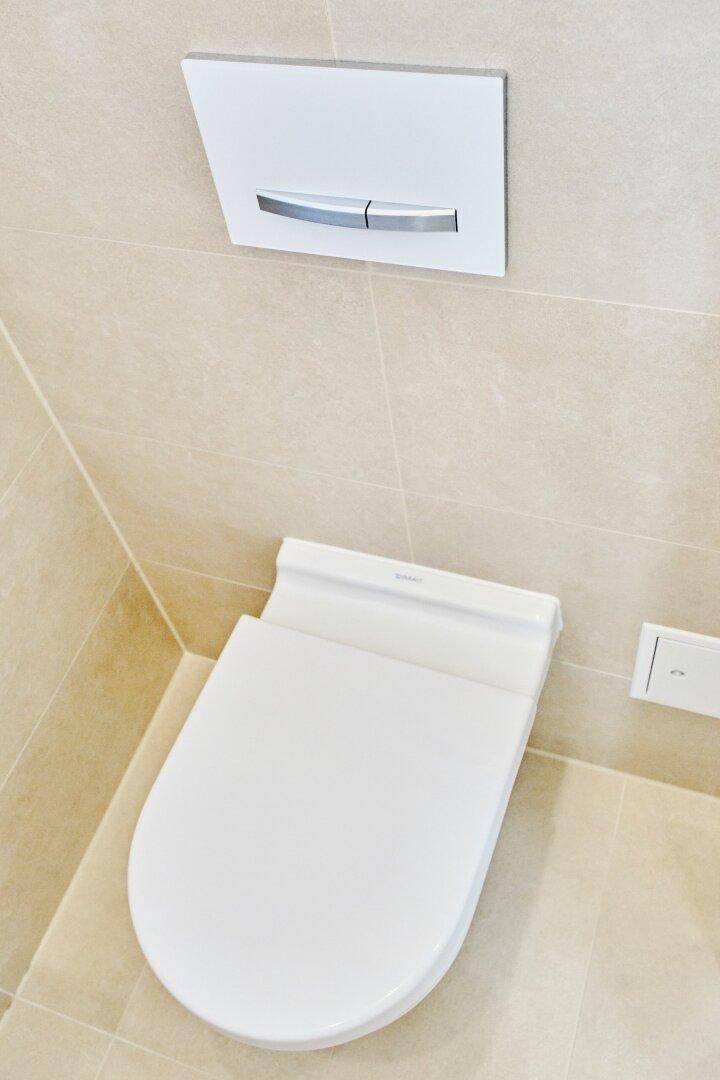 Wandhängende Toilette mit moderner Spültaste und beigefarbenen Fliesen, sauber und platzsparend.