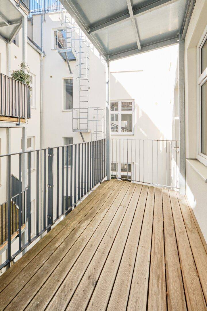 Heller Balkon mit Holzdielen und Metallgeländer, umgeben von der Fassade des Gebäudes.