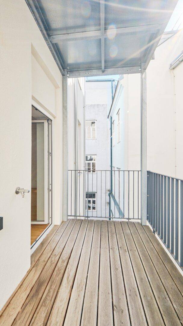 Schmaler Balkon mit Holzboden und Metallgeländer, der einen Blick auf die umliegenden Gebäude bietet.