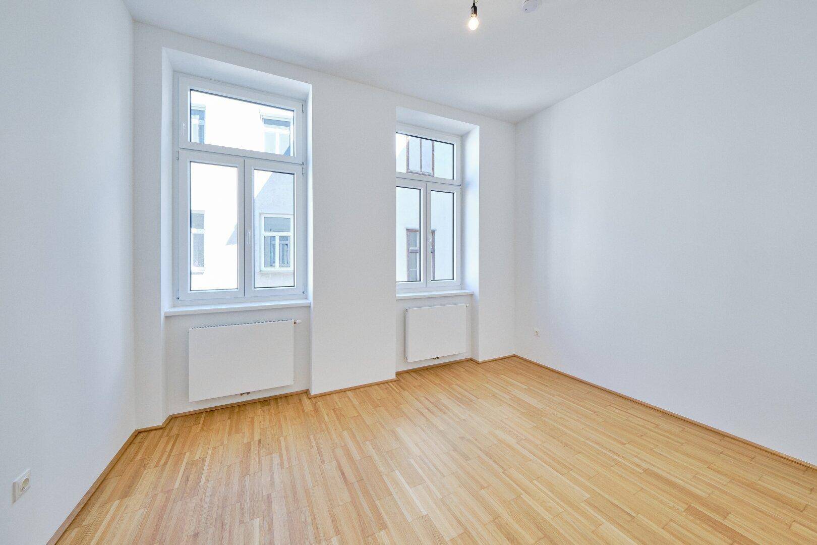 Geräumiges Zimmer mit zwei großen Fenstern und hellem Parkettboden, bietet viel Tageslicht.