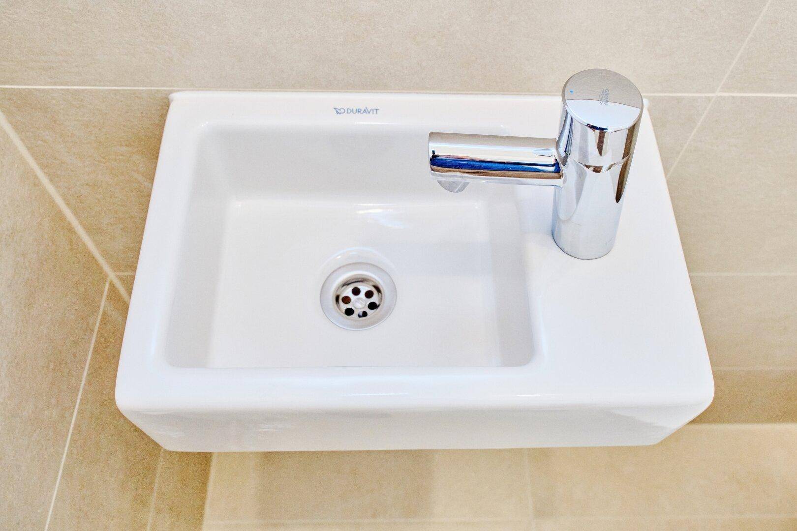 Kleines Waschbecken mit modernem Wasserhahn in einem hell gefliesten WC.