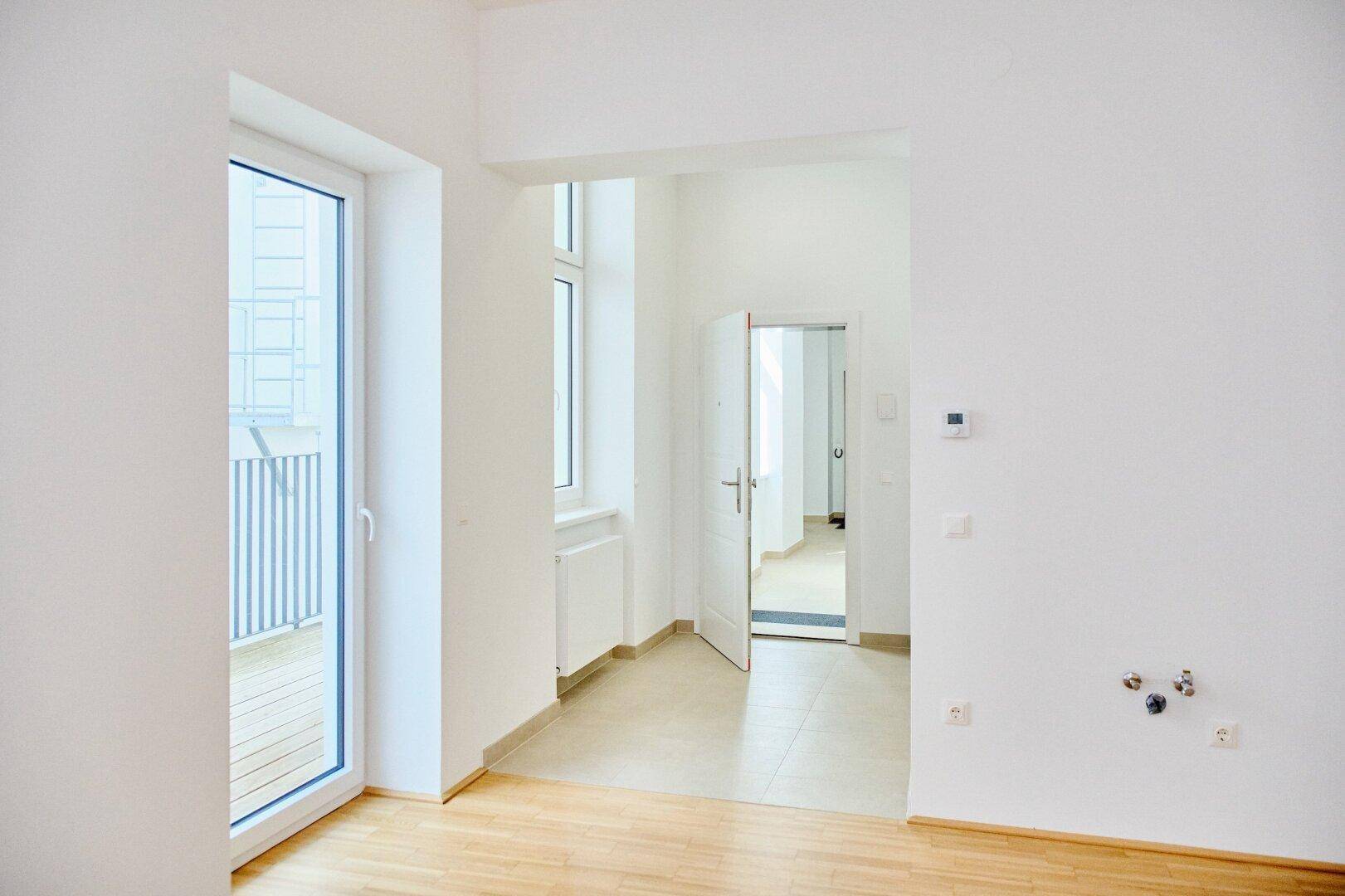 Heller Wohnbereich mit Parkettboden, Fenster und Zugang zum Balkon sowie weiteren Räumen.