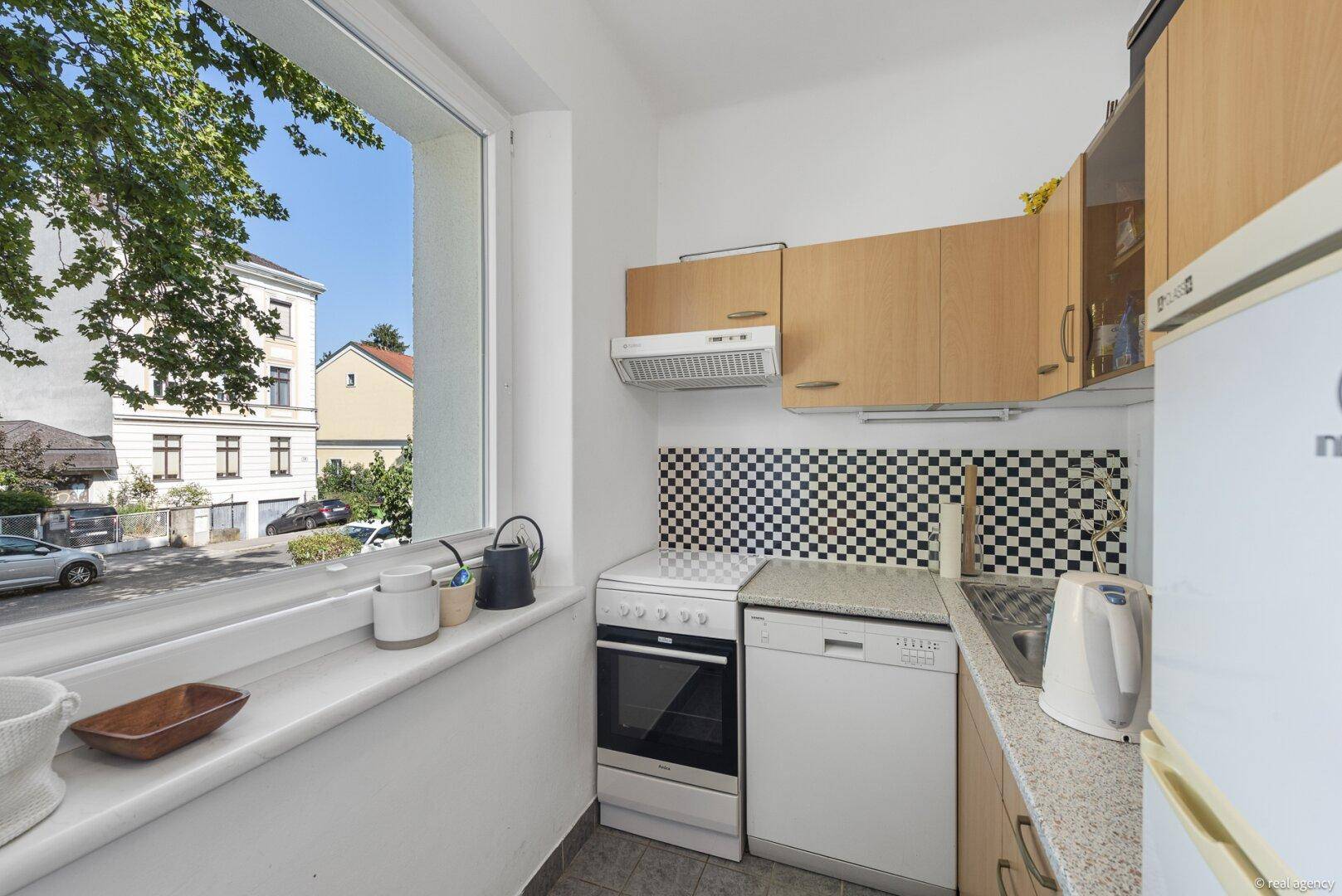 Kleine Küche mit Fenster, Einbaugeräten und einem schwarz-weiß karierten Fliesenspiegel.