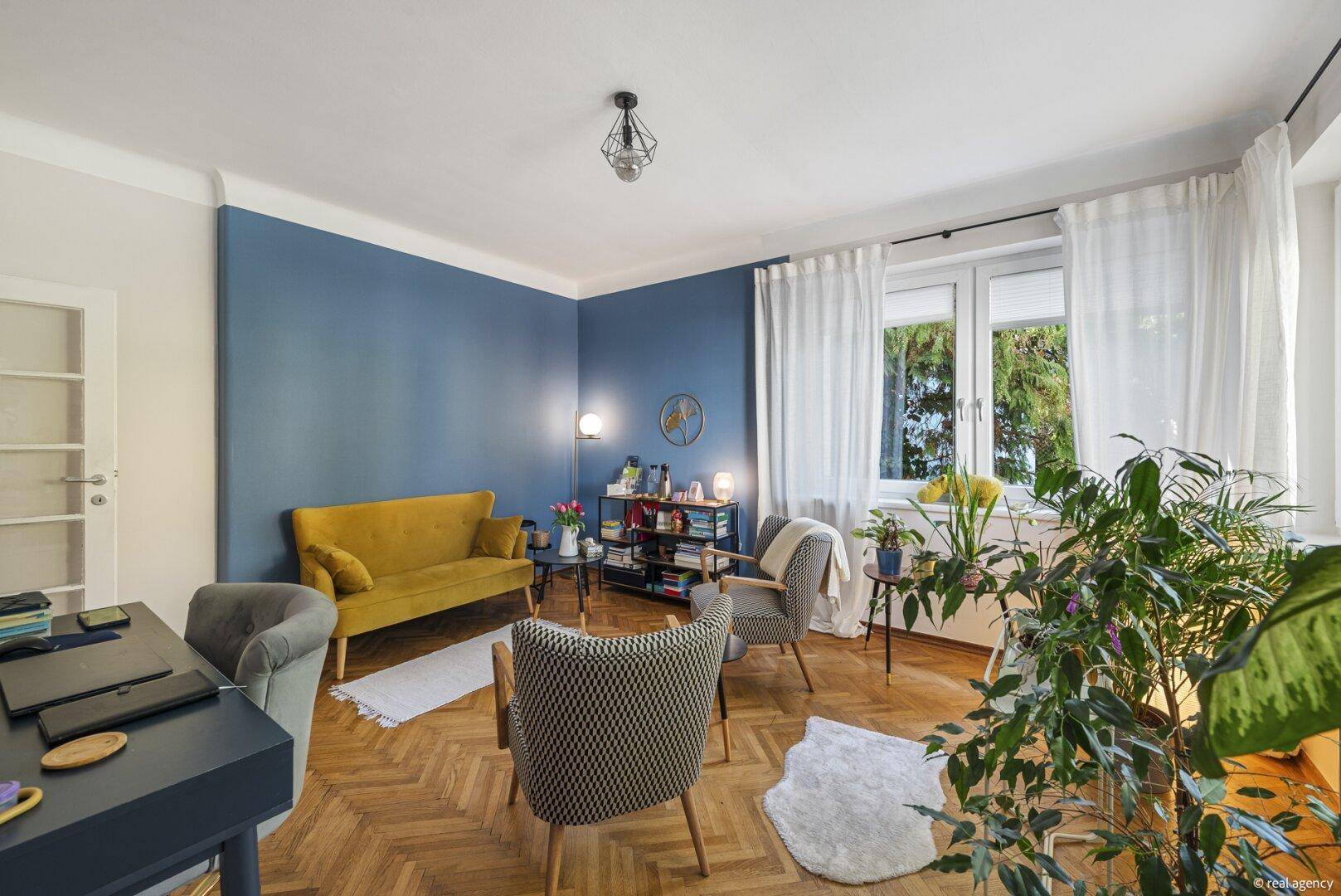 Gemütliches Wohnzimmer mit blauer Wand, gelbem Sofa und Blick ins Grüne durch große Fenster.
