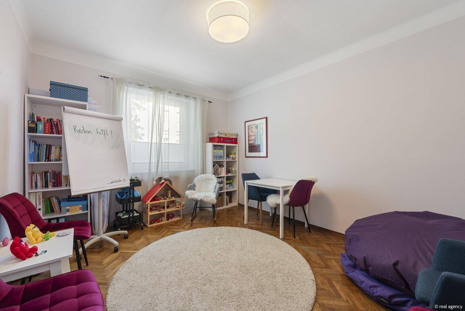 Helles Arbeitszimmer mit Parkettboden, Bücherregalen und einem runden Teppich in neutralen Farben.