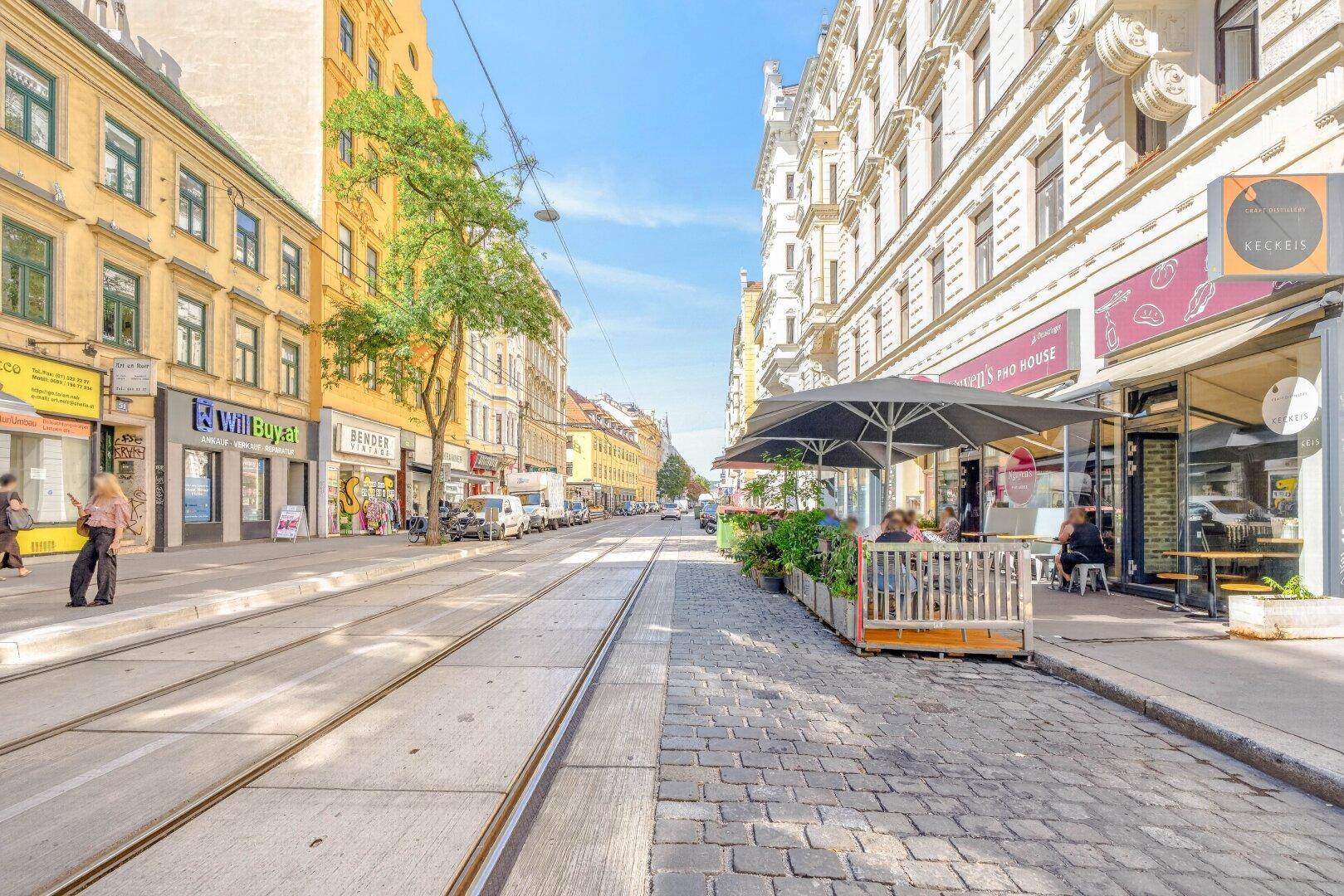 Eine breite Stadtstraße mit Kopfsteinpflaster, Straßenbahnschienen und Gebäuden mit Geschäften und Cafés.