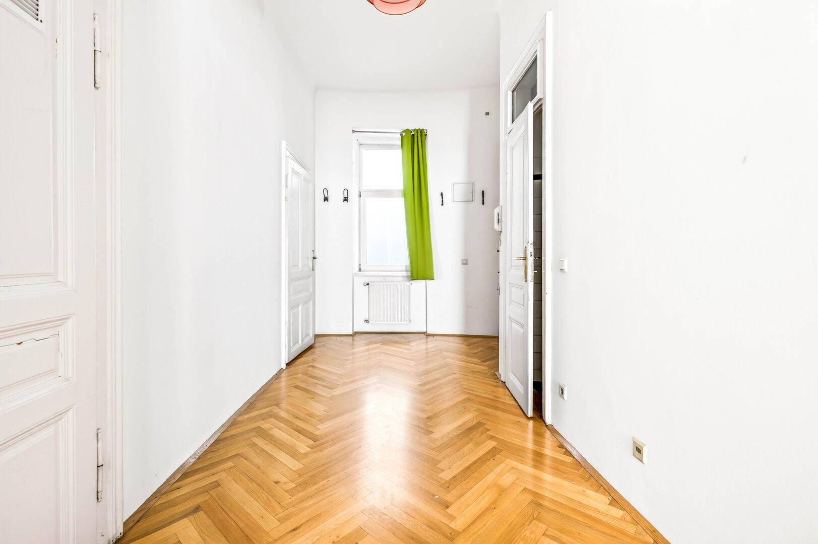 Ein heller Flur mit Fischgrätparkett, weißen Wänden und einem Fenster mit grünem Vorhang.