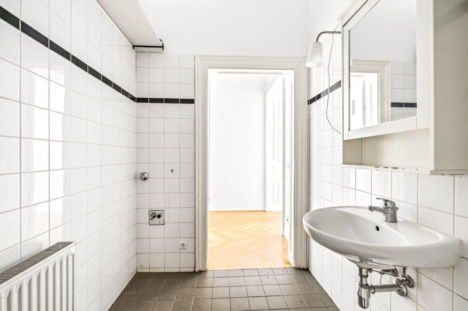 Ein Badezimmer mit weißen Fliesen, einem Waschbecken und einem Spiegelschrank, der zu einem anderen Raum führt.