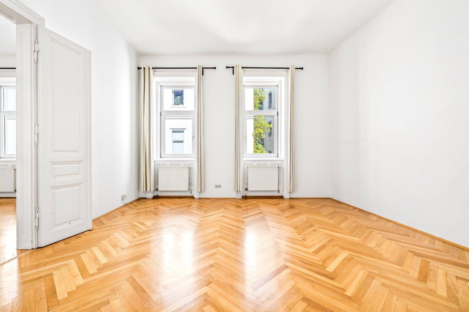 Ein heller Wohnraum mit Fischgrätparkett, weißen Wänden und zwei Fenstern mit Blick ins Grüne.