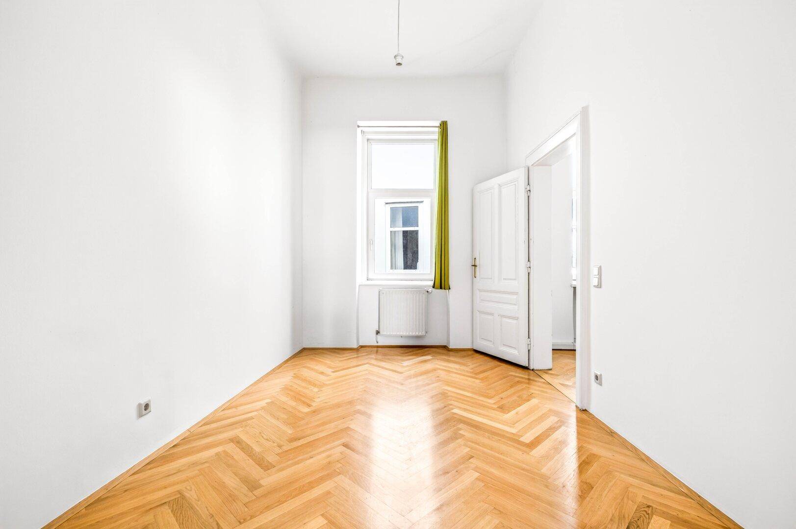 Ein helles Schlafzimmer mit Fischgrätparkett, weißen Wänden und einem Fenster mit grünem Vorhang.