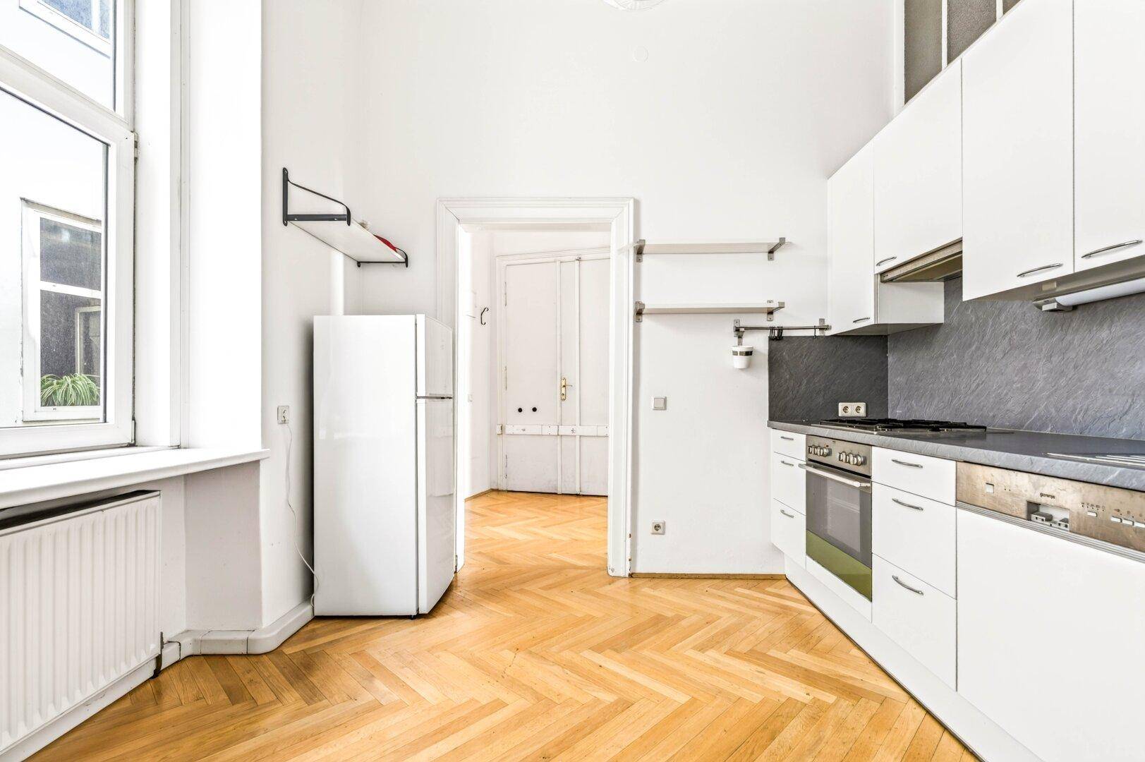 Eine helle Küche mit weißen Schränken, einem Kühlschrank und einem Fenster mit Blick nach draußen.