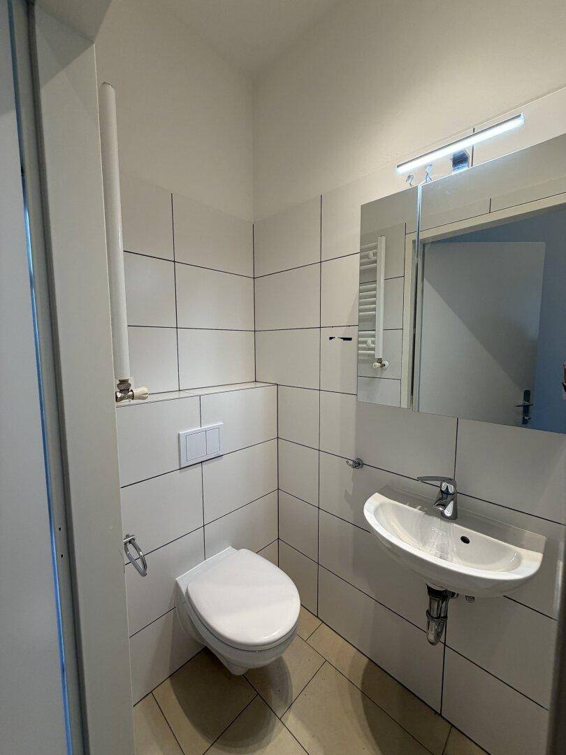 Modern gefliestes WC mit wandhängender Toilette, Waschbecken und Spiegel.