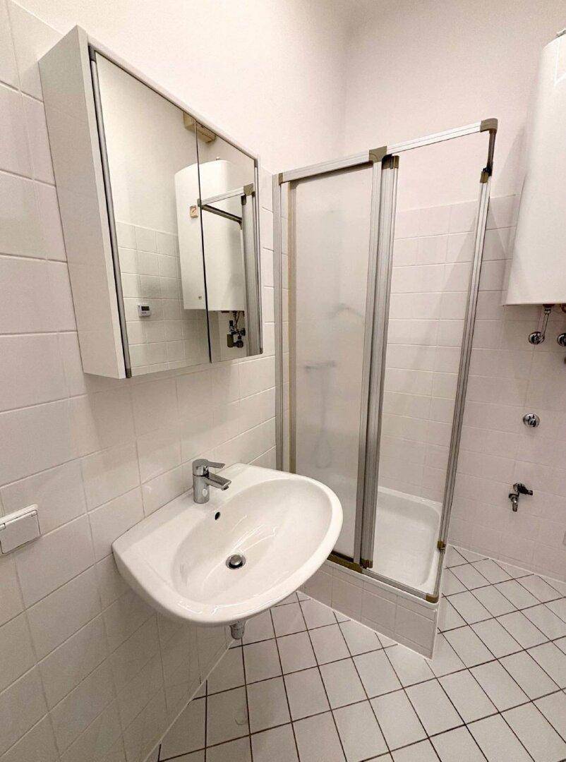 Funktionales Badezimmer mit weißen Fliesen, Waschbecken, Spiegelschrank und einer Duschkabine.