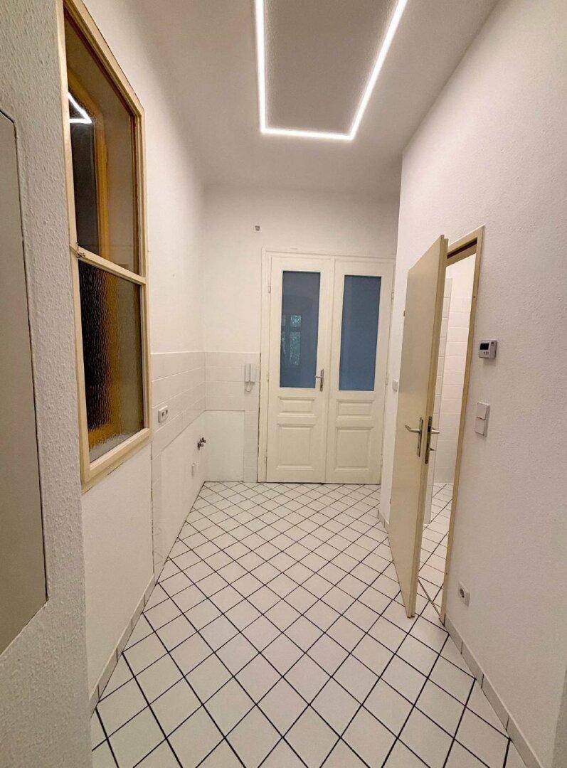 Heller Flur mit weißen Bodenfliesen, einer Doppeltür und einem Fenster, das Tageslicht spendet.