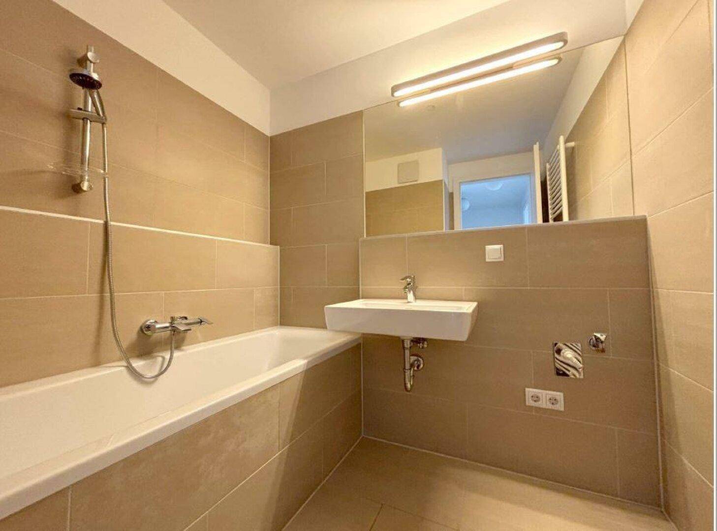 Modernes Badezimmer mit Badewanne, Duscharmatur, Waschbecken und großem Spiegel, gefliest in Beige.
