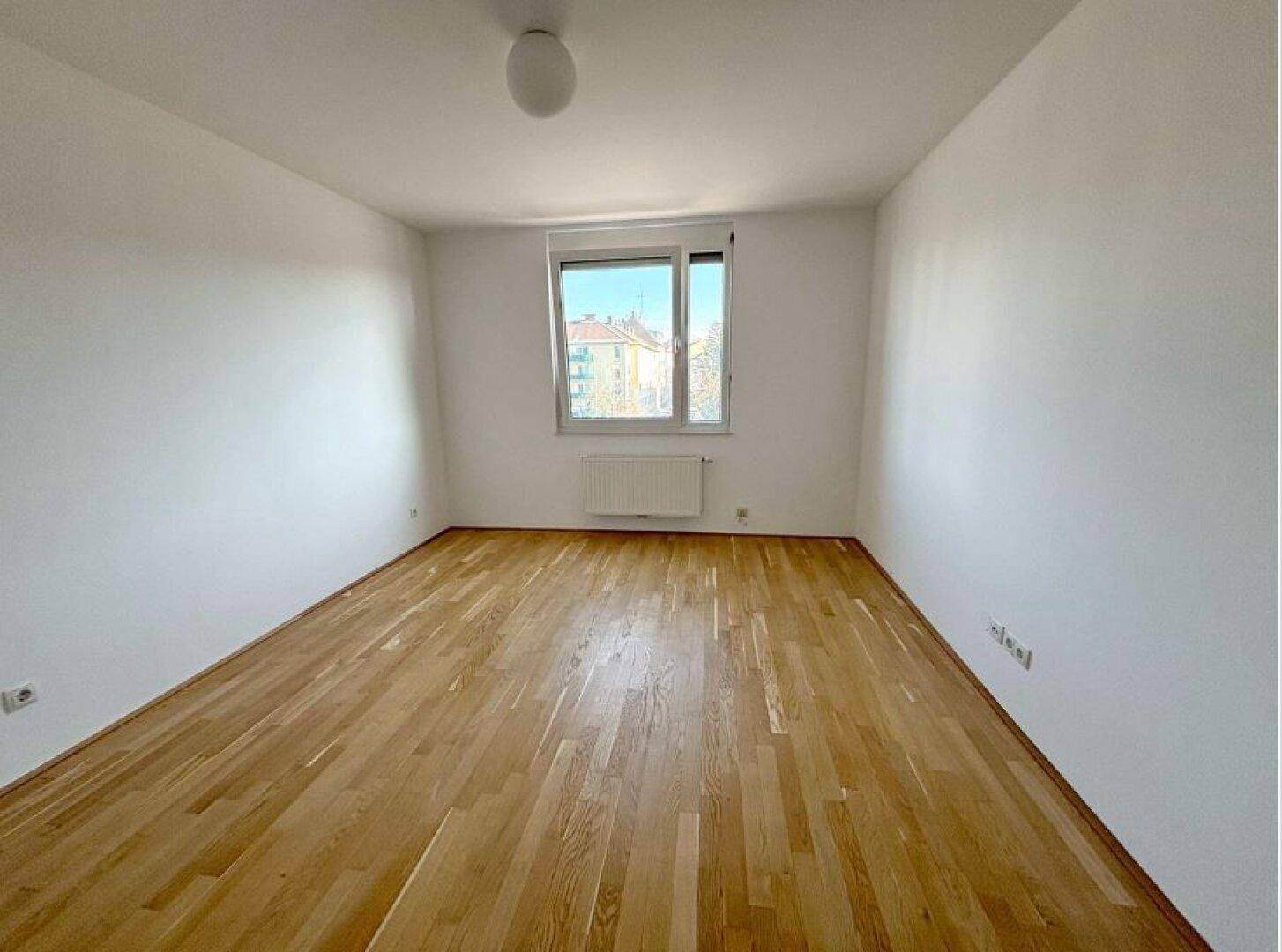 Leeres Zimmer mit hellem Holzparkettboden und einem Fenster, das viel Tageslicht hereinlässt.