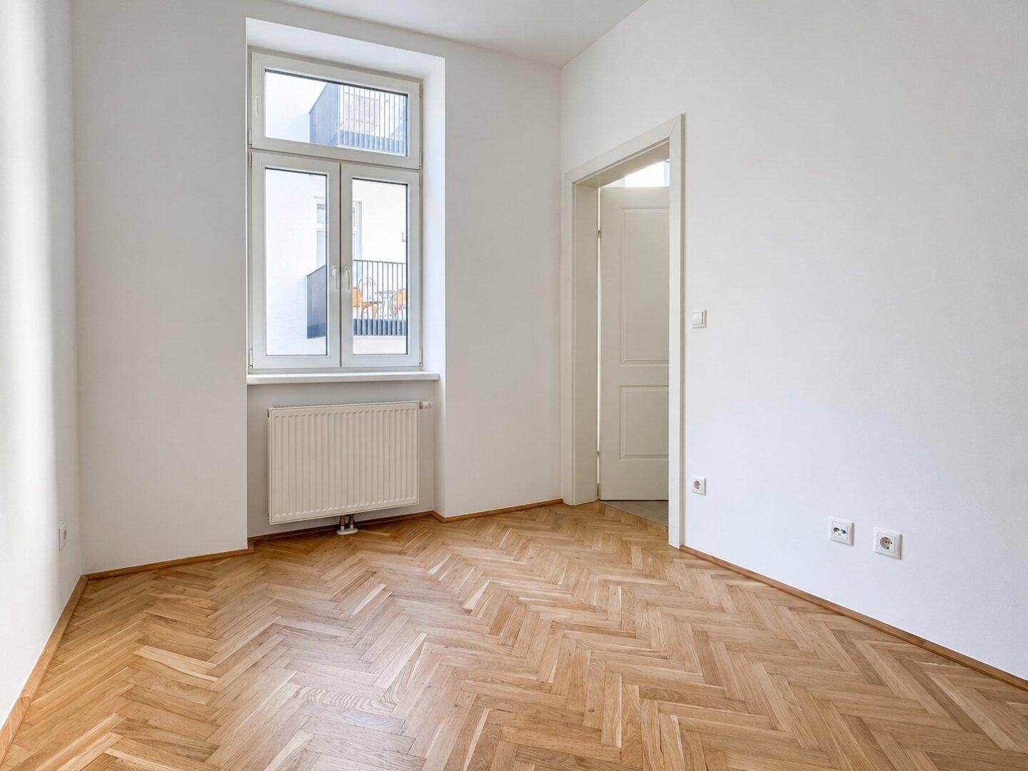 Freundliches Zimmer mit Fischgrätparkett und einem Fenster, das auf einen Balkon blickt.