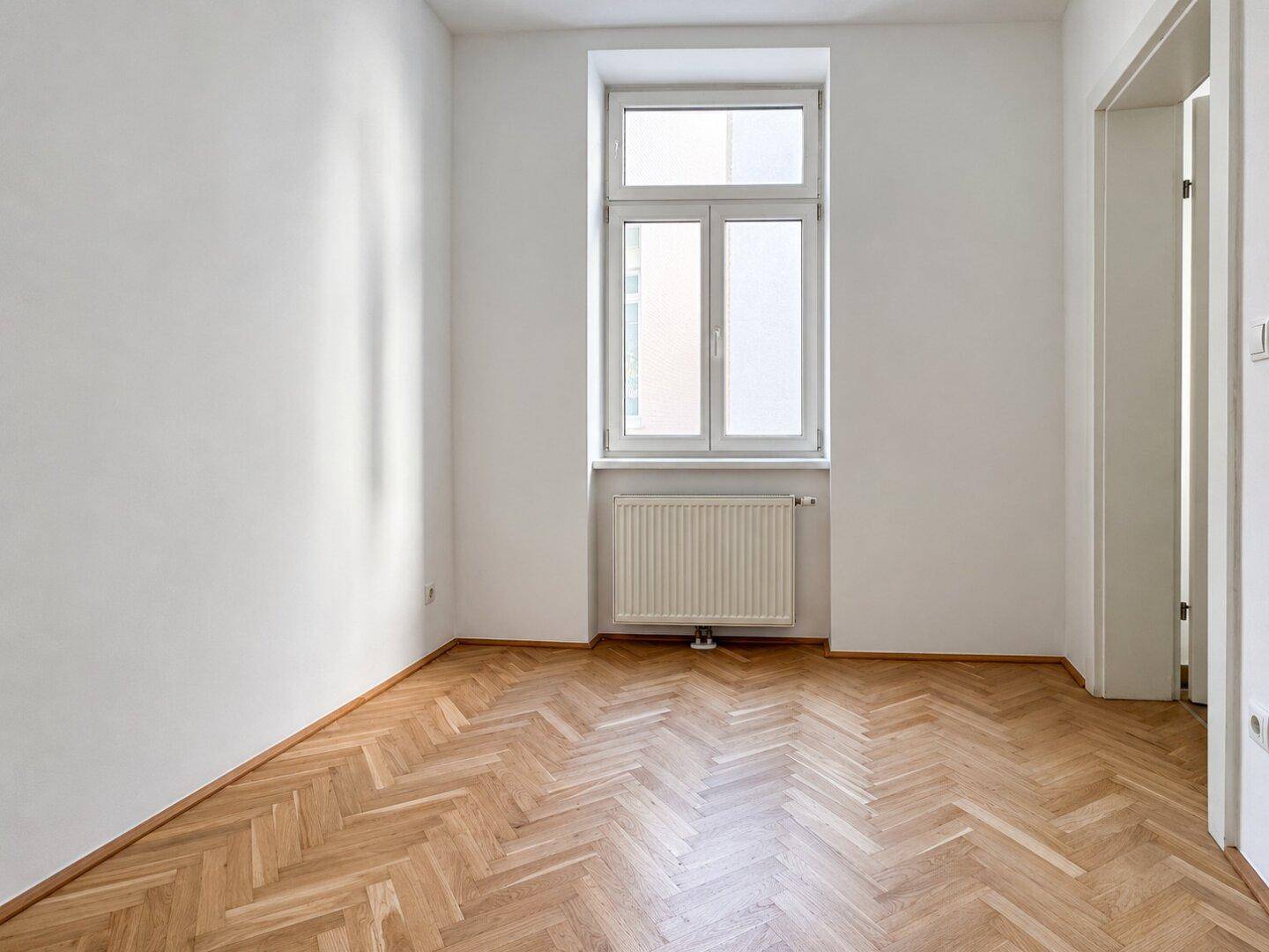 Leeres Zimmer mit hellem Fischgrätparkett und einem großen Fenster, bereit zum Einrichten.