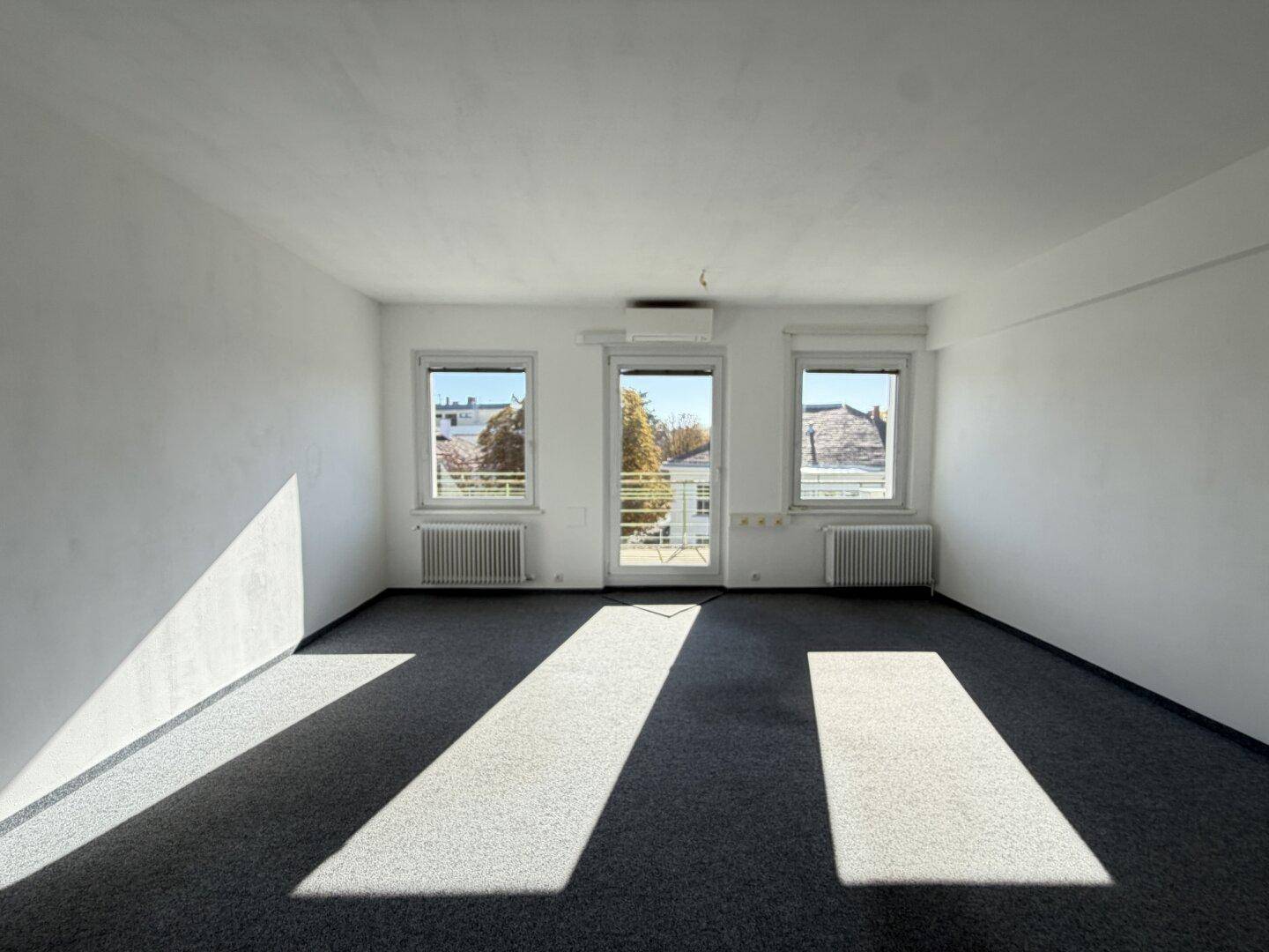 Heller, leerer Raum mit grauem Teppichboden und viel natürlichem Licht durch die Fenster.