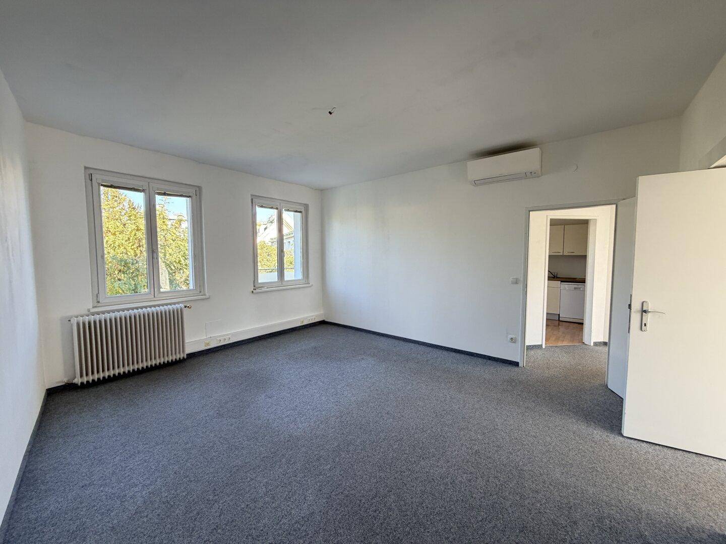 Geräumiger, leerer Raum mit grauem Teppichboden, Fenstern und Zugang zu einem Nebenraum.