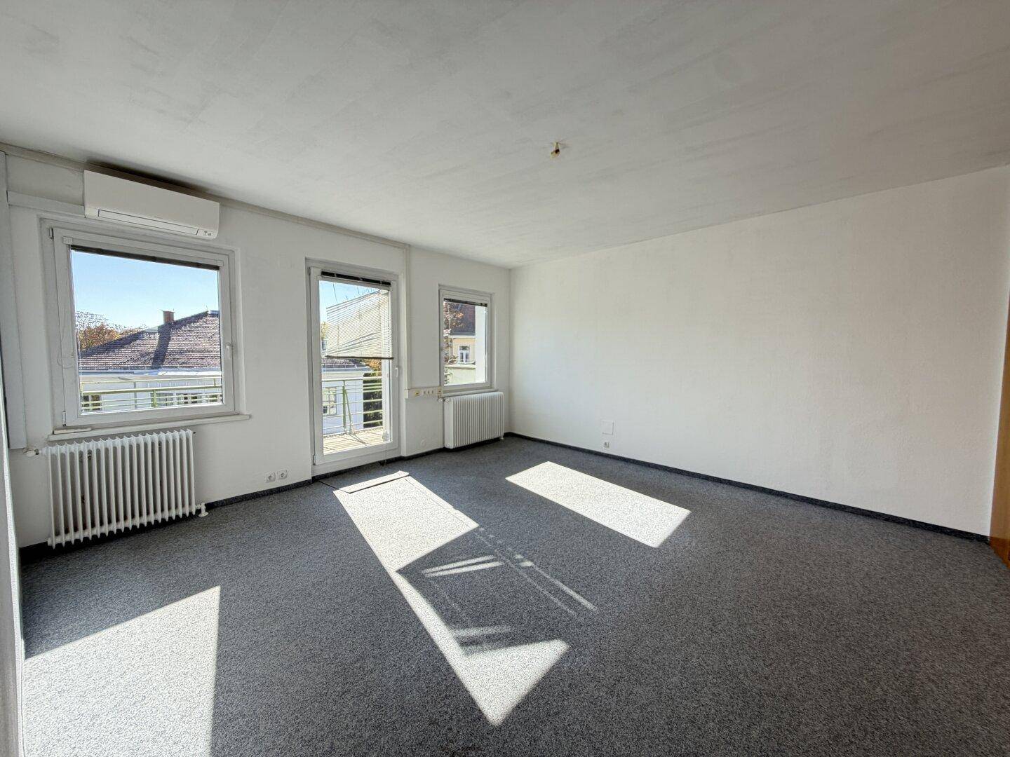 Heller Raum mit grauem Teppichboden, Fenstern und viel Sonnenlicht, das den Boden erhellt.