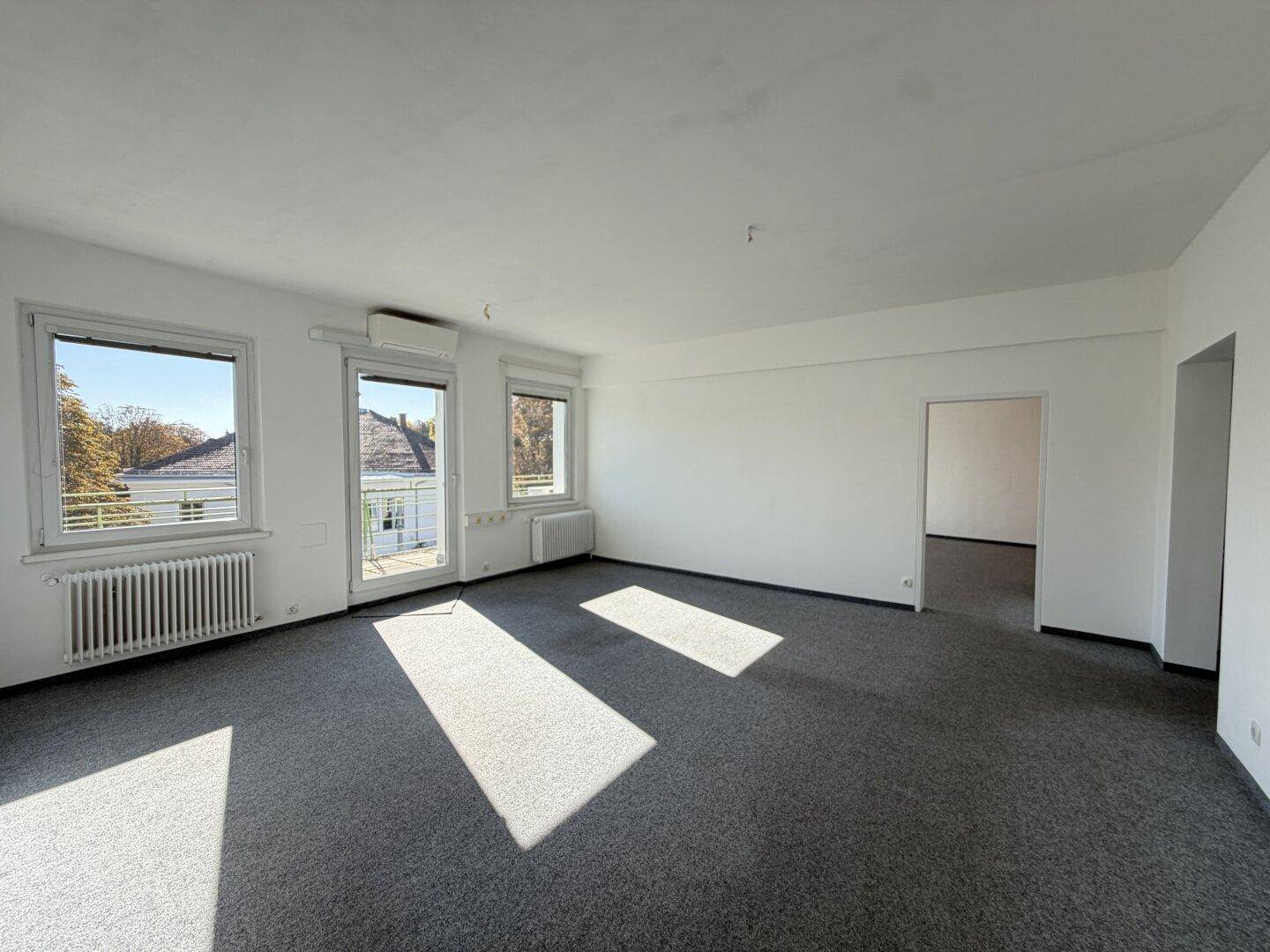 Geräumiger, heller Raum mit grauem Teppichboden, Fenstern und offenem Zugang zu weiteren Bereichen.