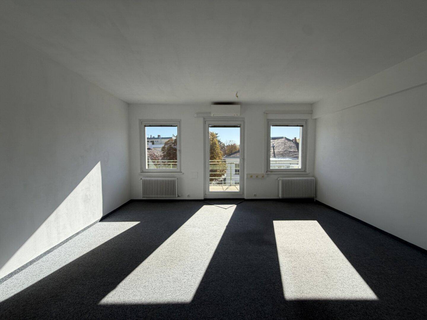 Heller, leerer Raum mit grauem Teppichboden und viel natürlichem Licht durch die Fenster.