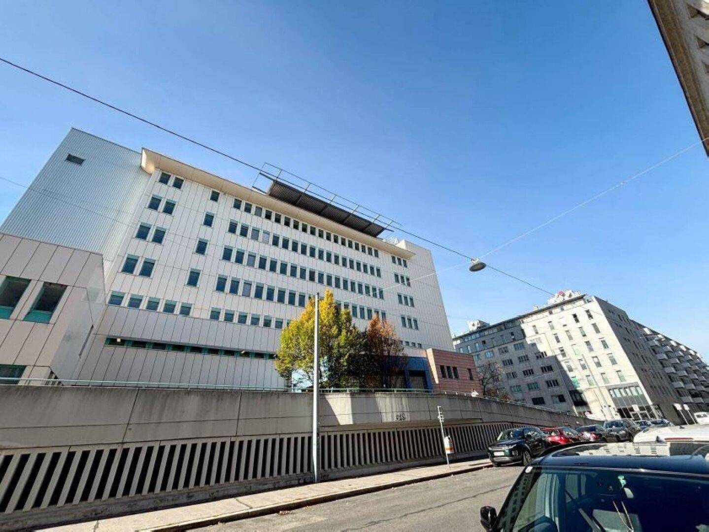 Moderne Krankenhausfassade mit vielen Fenstern und einer Zufahrtsrampe unter blauem Himmel.