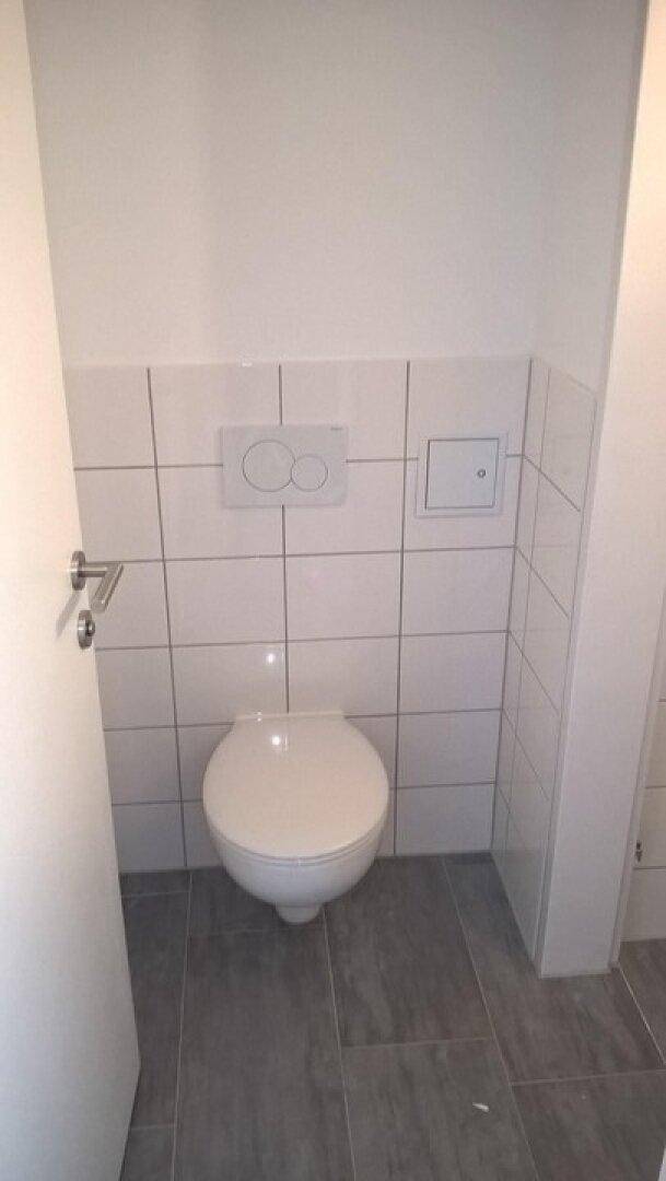Separates WC mit wandhängender Toilette und hellen Fliesen an den Wänden und auf dem Boden.