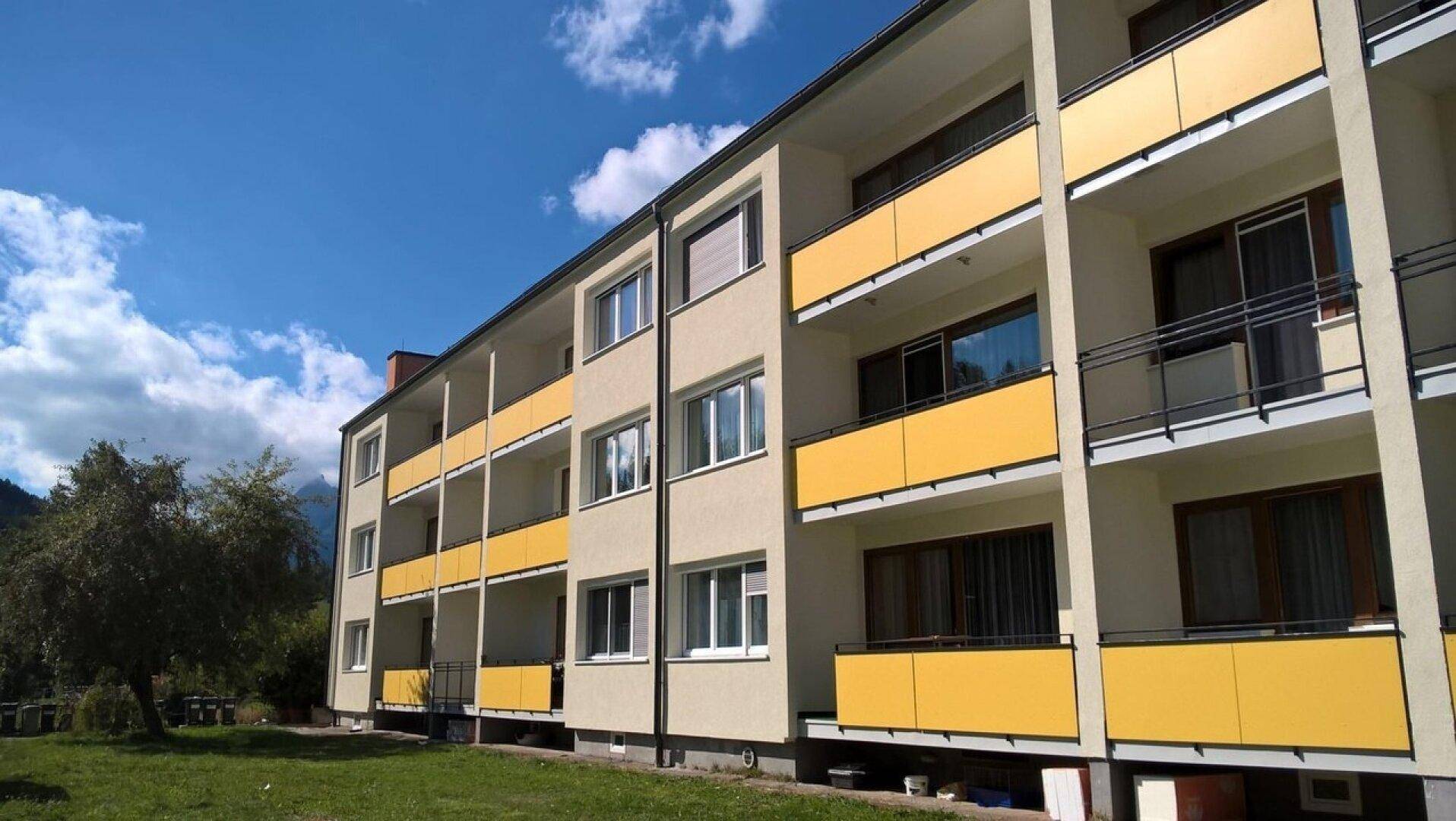 Modernes Mehrfamilienhaus mit gelben Balkonbrüstungen und gepflegter Grünfläche unter blauem Himmel.
