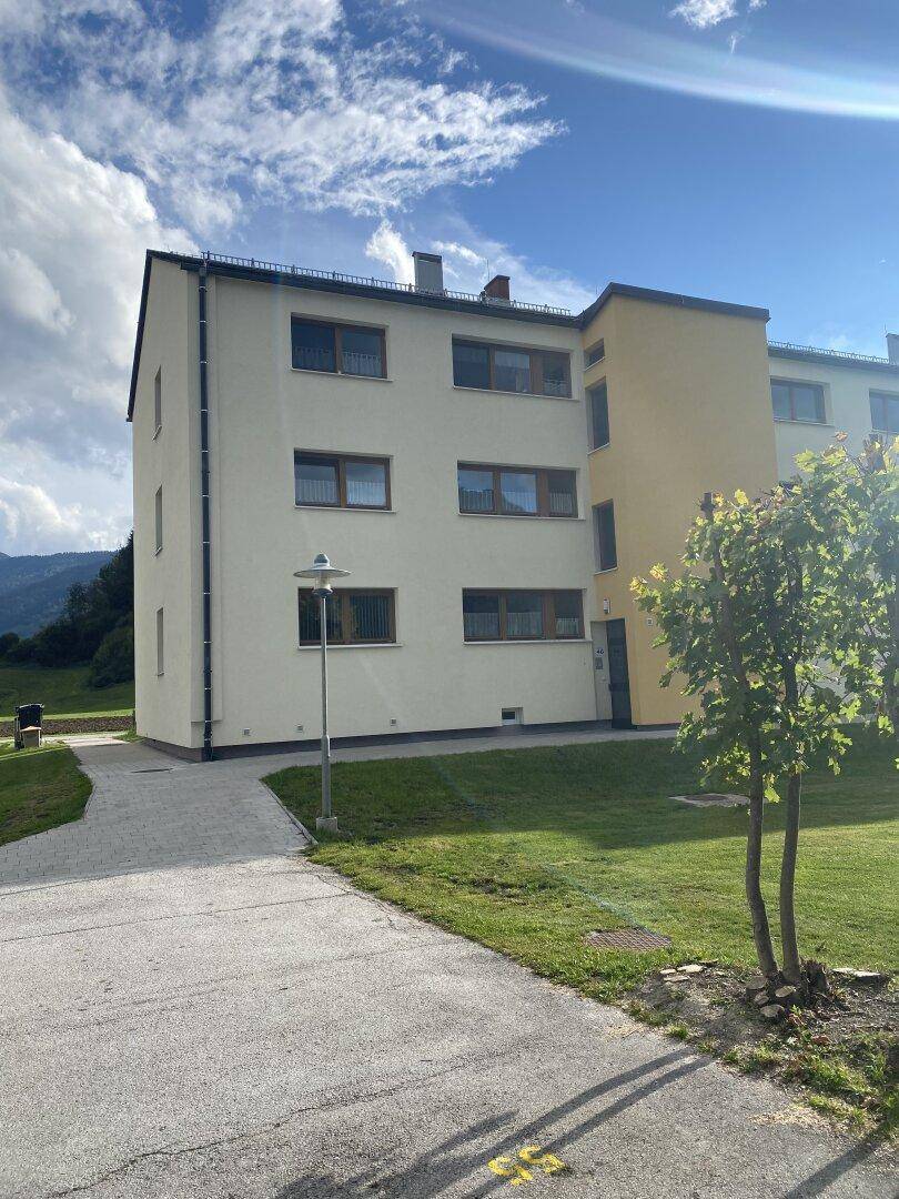Seitenansicht eines modernen Mehrfamilienhauses mit gepflegter Grünfläche und beeindruckender Bergkulisse im Hintergrund.