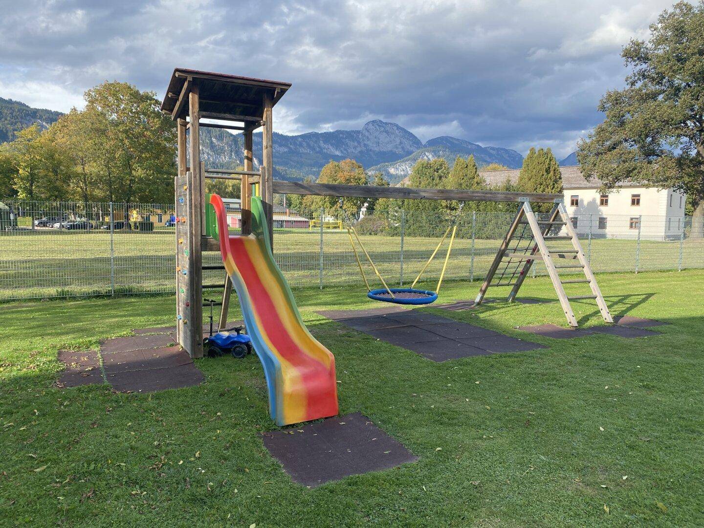 Kinderspielplatz mit Rutsche und Schaukel auf einer grünen Wiese vor einer malerischen Berglandschaft.