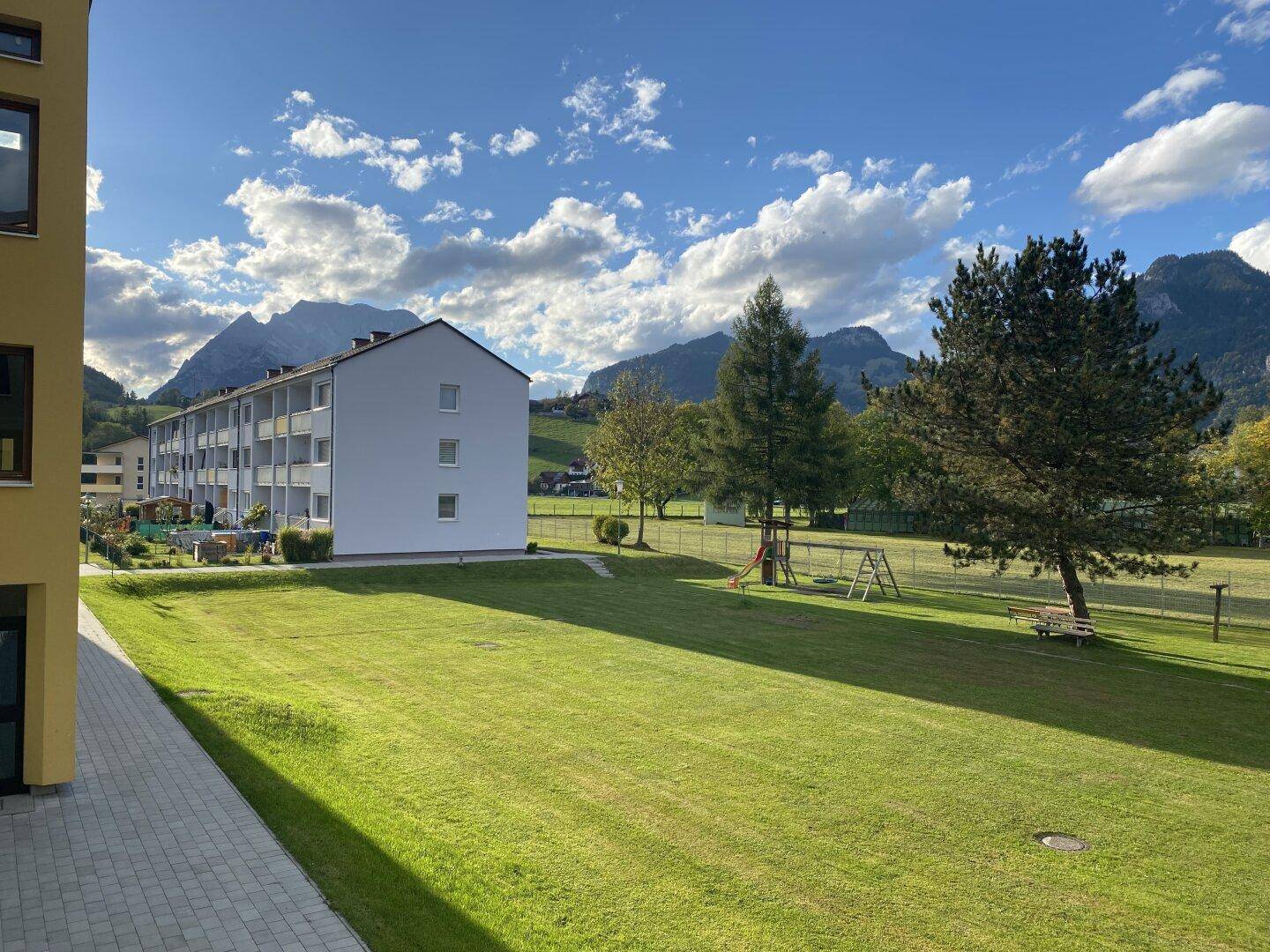 Weitläufige Grünfläche mit Spielplatz und Wohngebäuden im Hintergrund, umgeben von einer Bergkulisse.