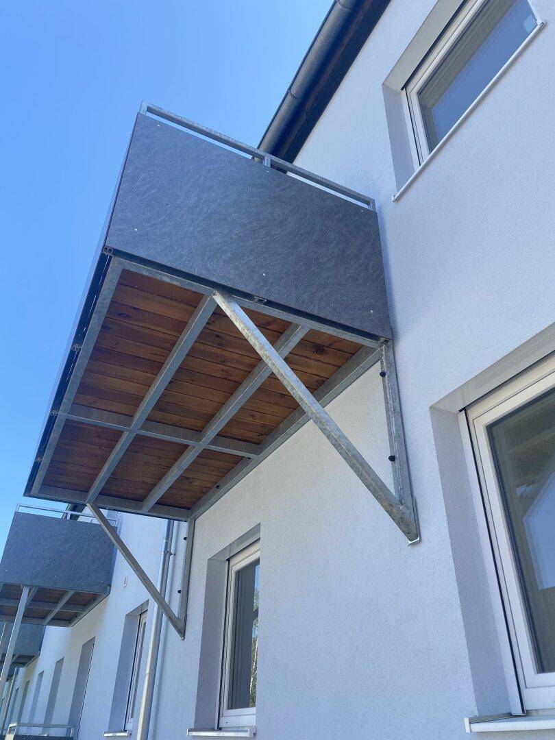 Außenansicht eines modernen Balkons mit Holzunterseite und Metallkonstruktion an einer weißen Fassade.