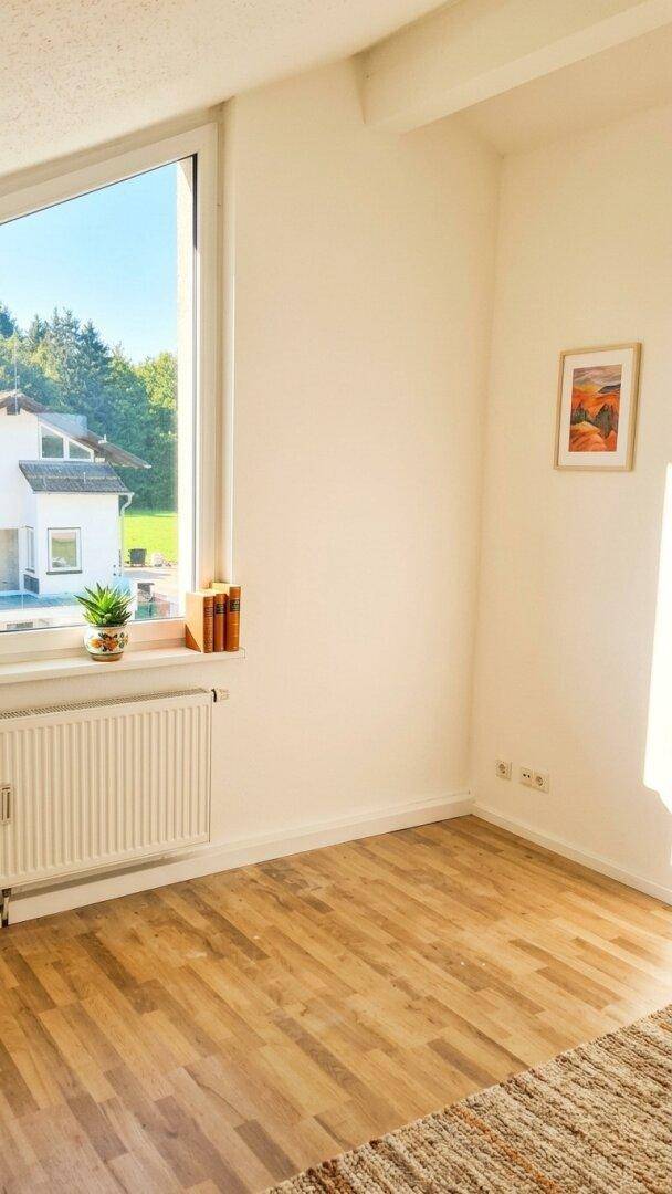 Heller Raum mit großem Fenster, Holzboden und Blick auf die umliegende Bebauung und Bäume.