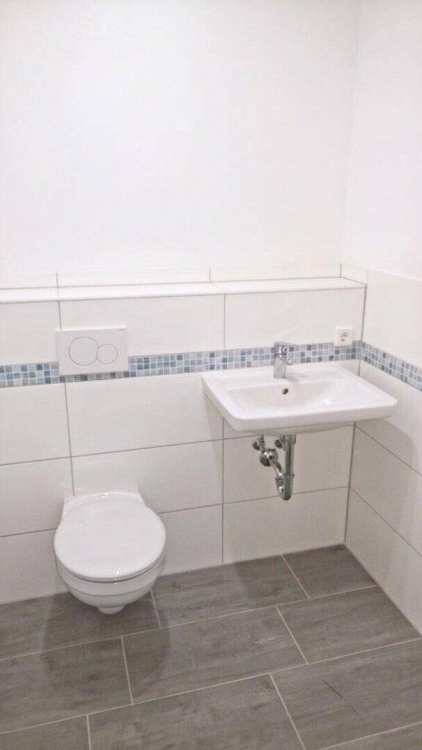 Helles WC mit Wand-WC, Waschbecken und weißen Fliesen mit blauer Mosaikbordüre.