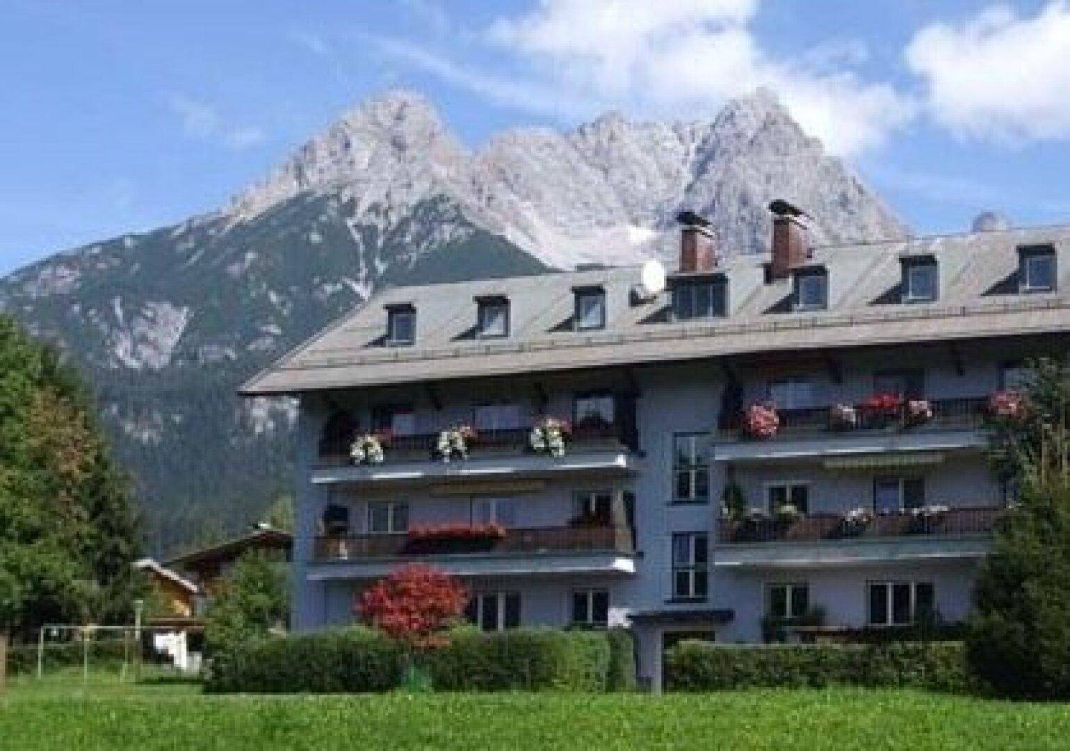 Mehrstöckiges Apartmentgebäude mit Balkonen und Blumenkästen vor einer beeindruckenden Bergkulisse.