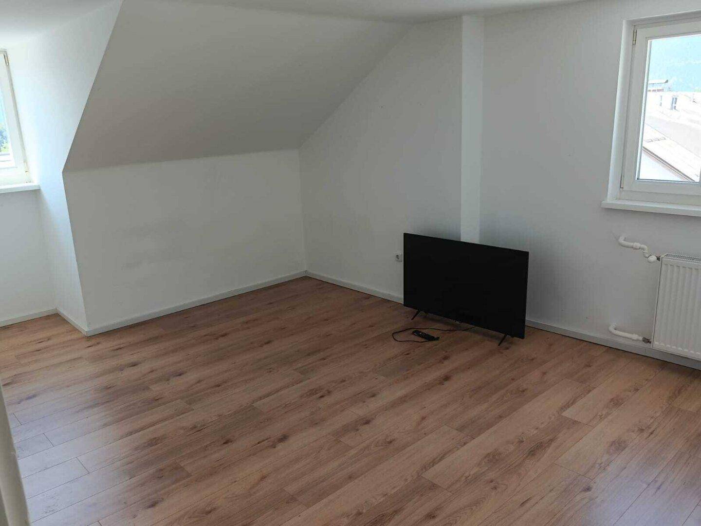 Helles Zimmer mit Dachschräge, Laminatboden und einem Fenster, ausgestattet mit einem Fernseher.