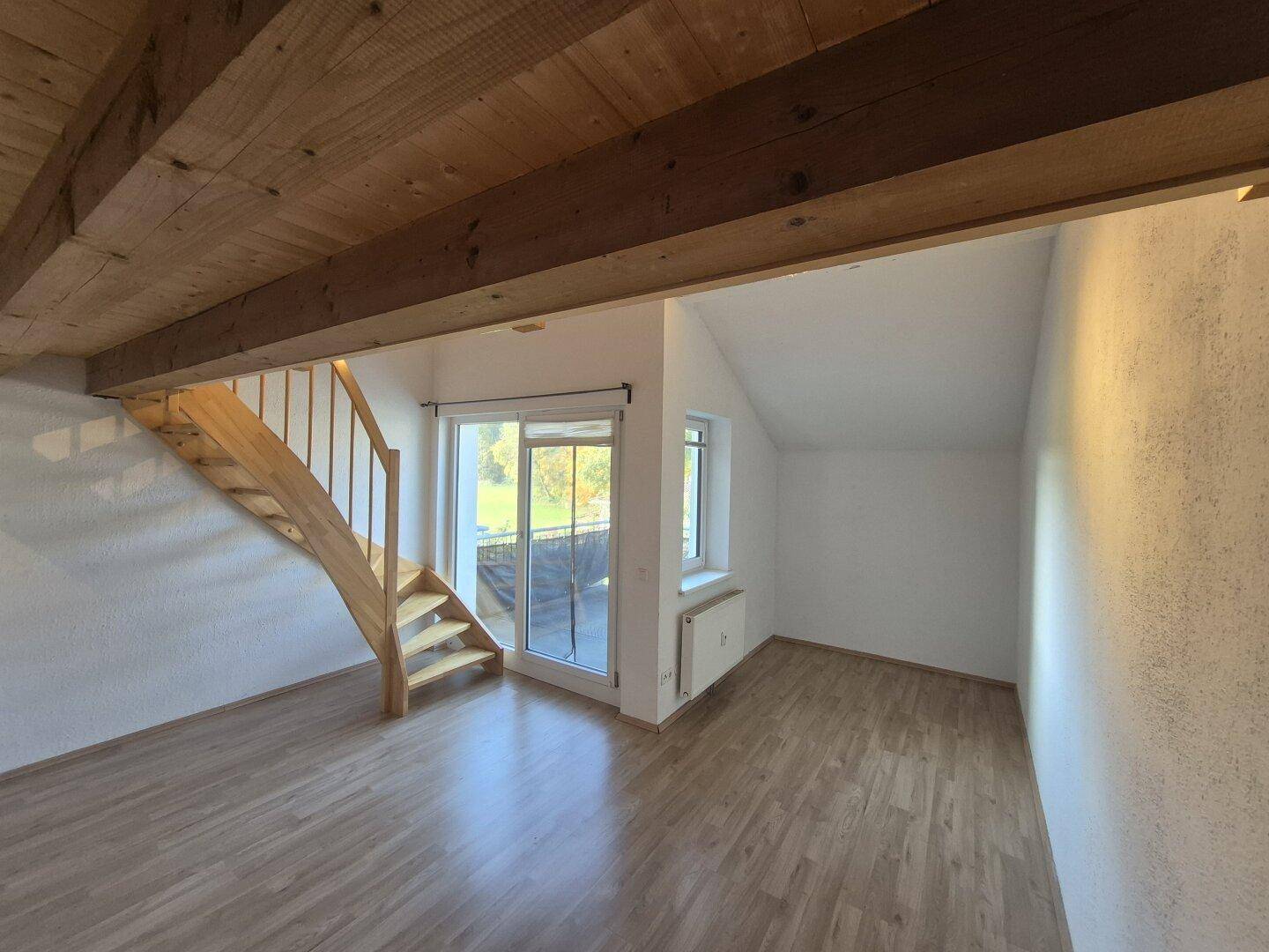Heller Wohnbereich mit Holzbalken, Laminatboden und Zugang zu einem Balkon.