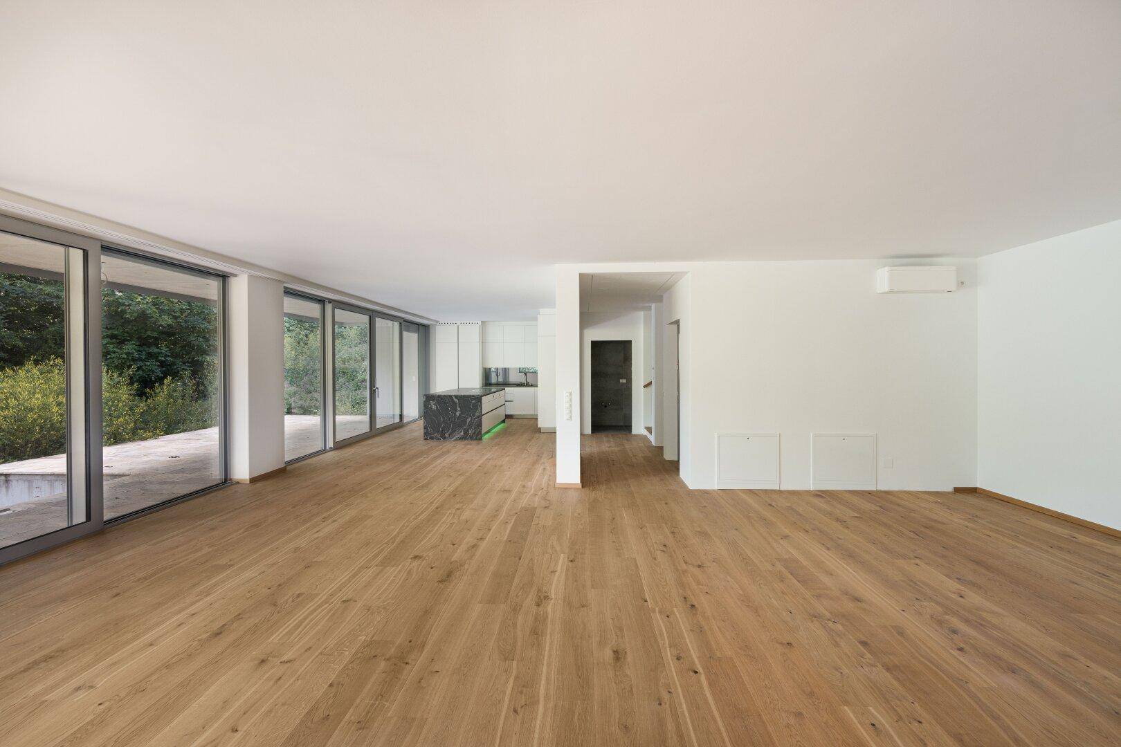 Offener Wohnbereich mit Holzboden, großen Fenstern und Blick auf die moderne Küche.