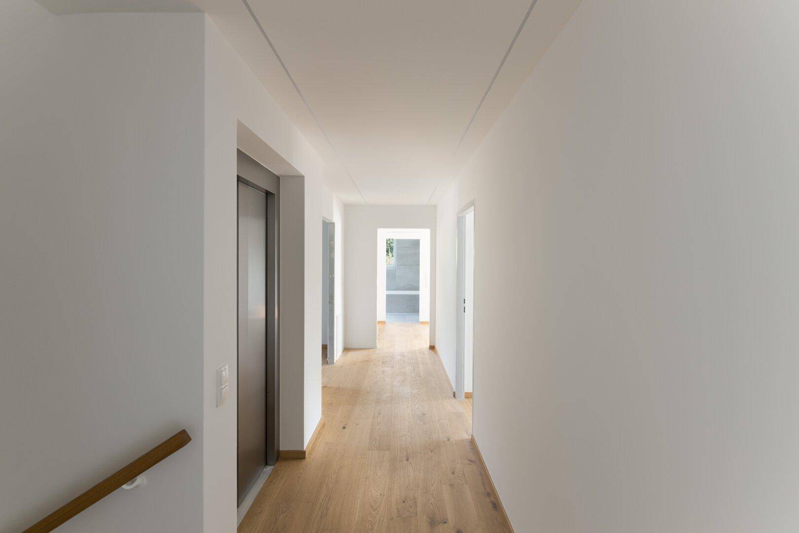 Langer, heller Flur mit Holzboden, integrierter Deckenbeleuchtung und Aufzugzugang.