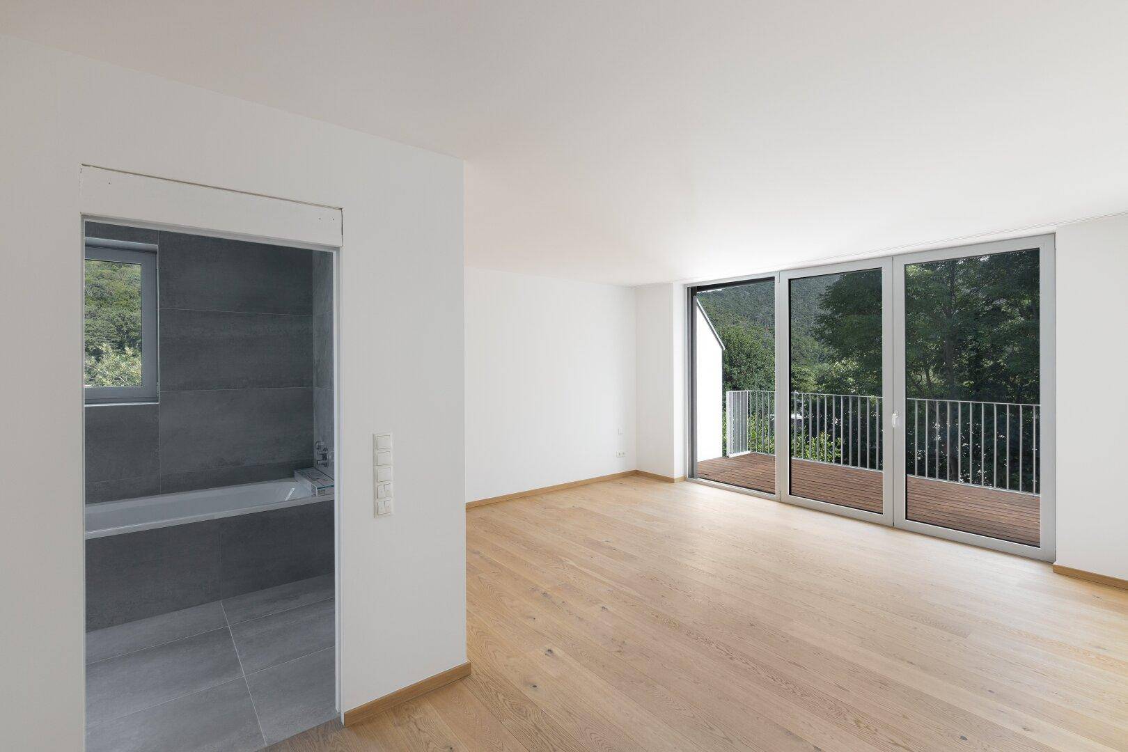 Helles Schlafzimmer mit Holzboden, Balkonzugang und angrenzendem Badezimmer.