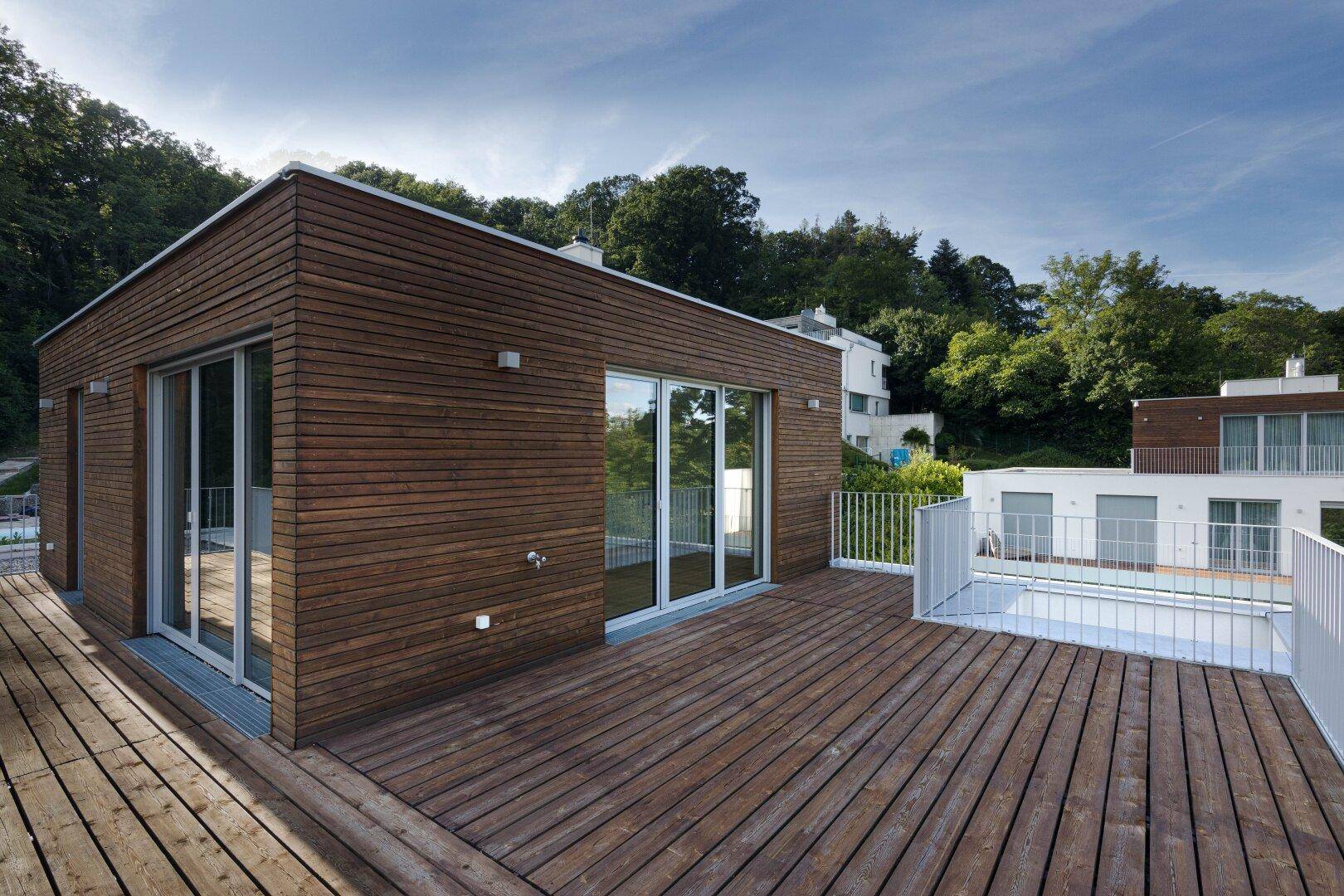 Moderne Holzfassade mit großer Terrasse und Blick auf weitere Gebäude in grüner Umgebung.
