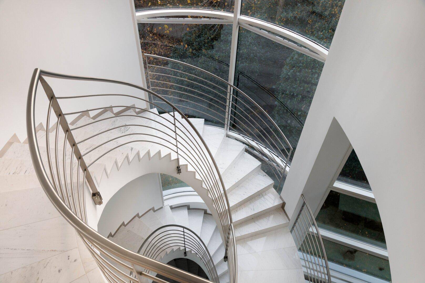 Elegante Wendeltreppe mit Marmorstufen und Edelstahlgeländer unter einem Glasdach.