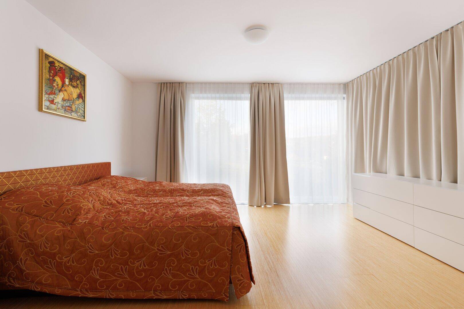 Schlafzimmer mit großen Fenstern, hellen Vorhängen und modernem Holzboden.