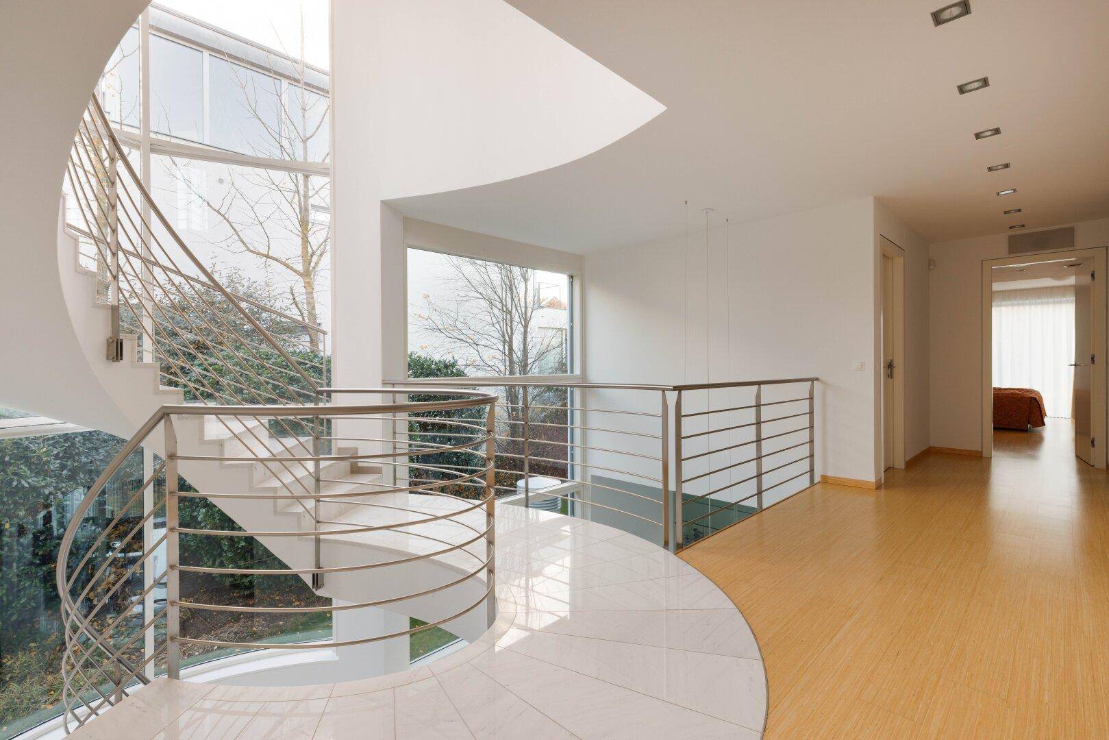Elegante Wendeltreppe mit Marmorstufen und Edelstahlgeländer in einem lichtdurchfluteten Flur.