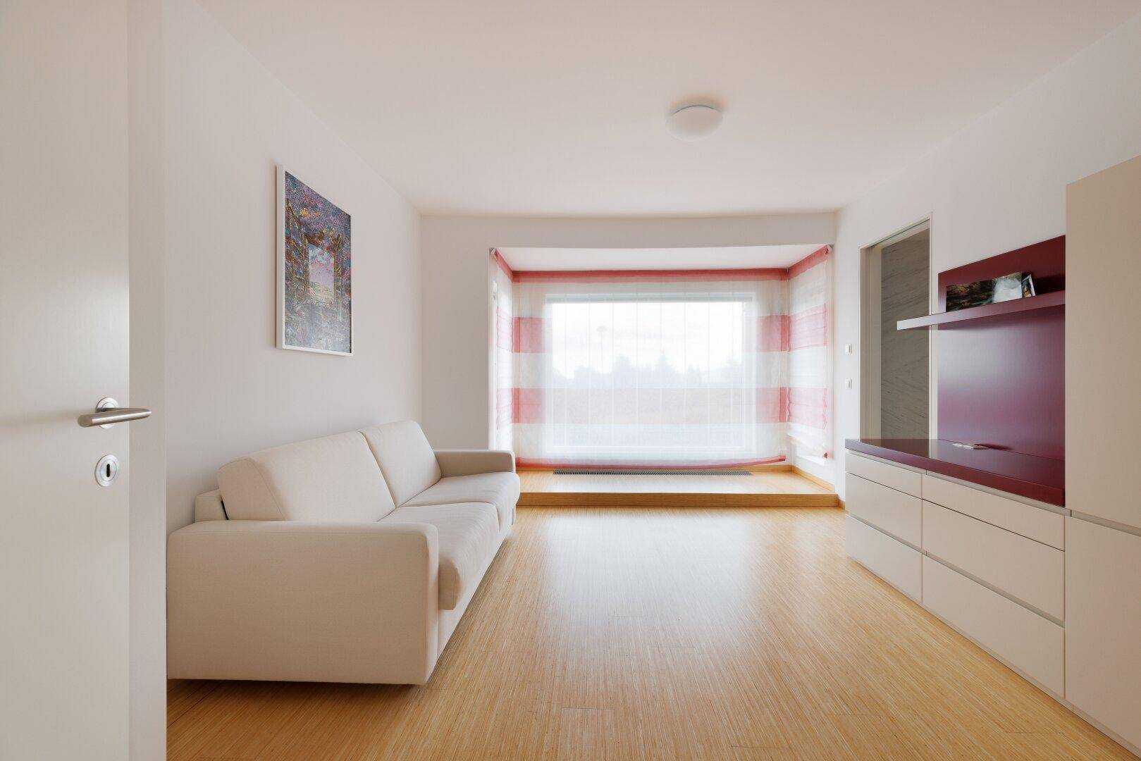 Gemütliches Wohnzimmer mit hellem Holzboden und einem großen Fenster mit Blick ins Grüne.