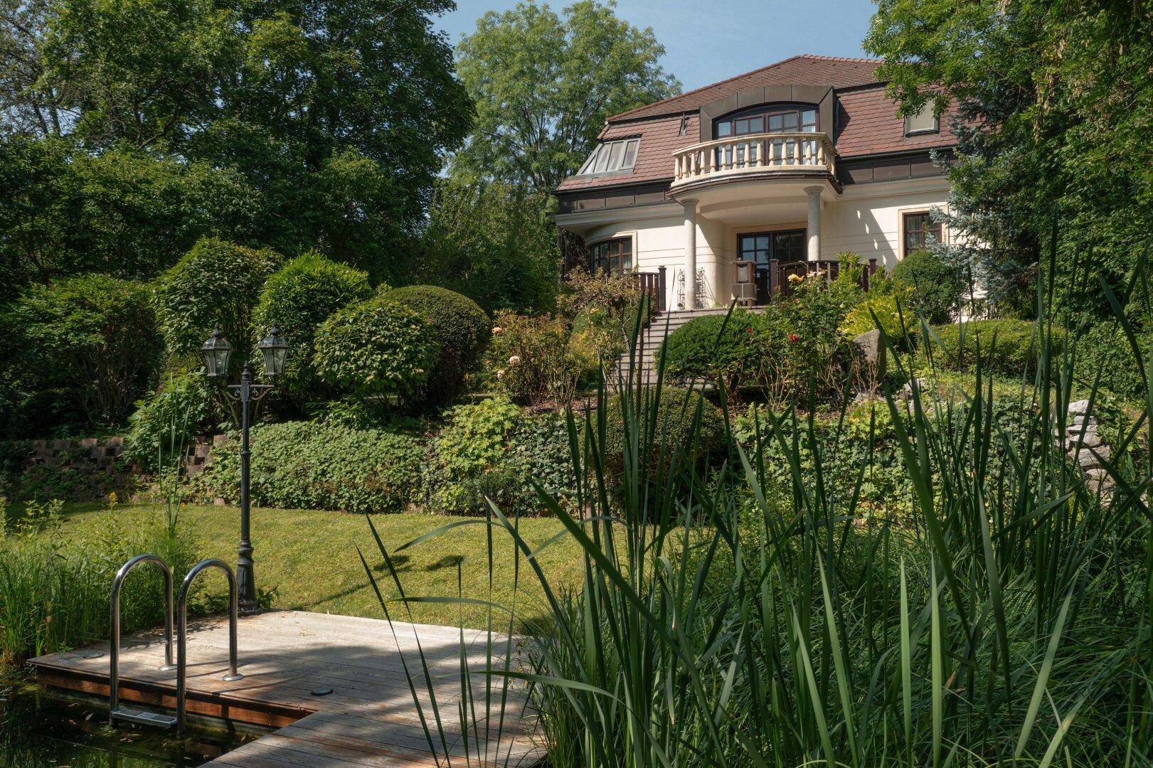 Herrschaftliche Villa mit gepflegtem Garten, Teich und üppiger Vegetation an einem sonnigen Tag.