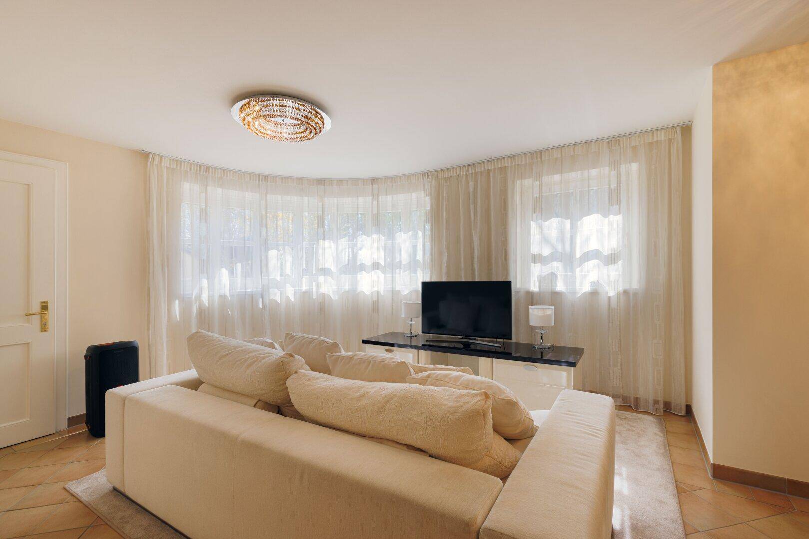 Helles Wohnzimmer mit beigem Sofa, Fernseher und großen Fenstern mit transparenten Vorhängen.